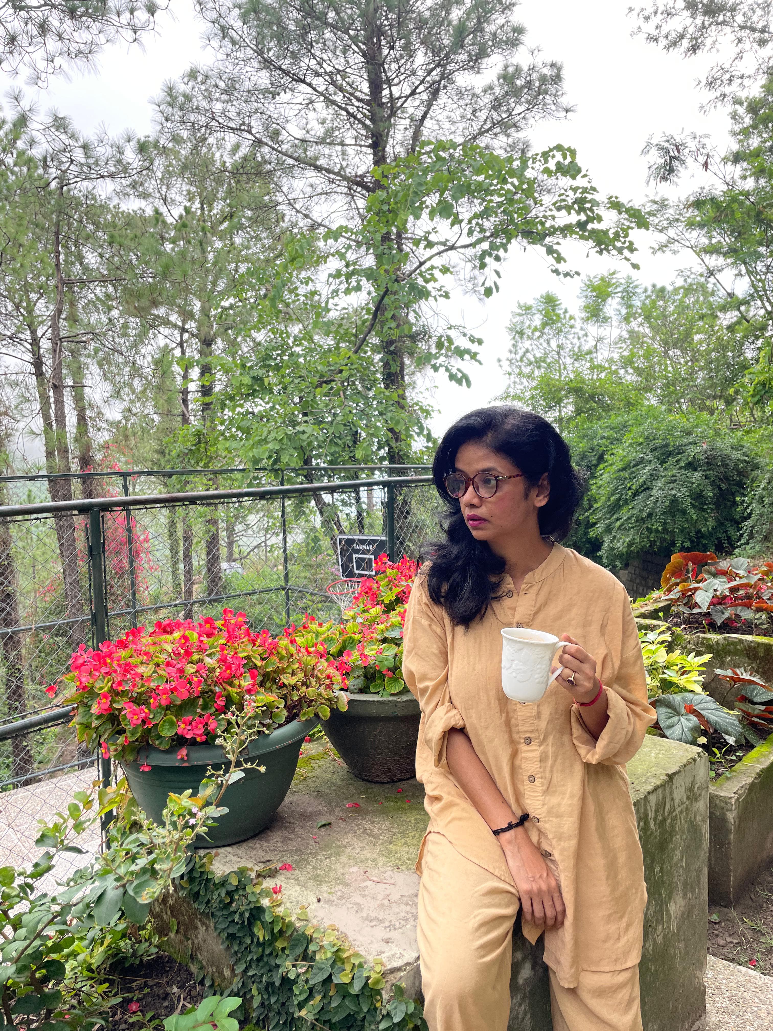 A female customer wearing a Paizlie beige gold silk loungewear set, taking photo outdoors in a beautiful hill resort, early in the  morning, while drinking her coffee.
